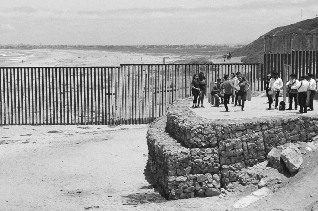 A group of people stand near a metal border fence that extends into the ocean, with stone walls and a sandy beach in the foreground, and distant cityscape visible across the water.