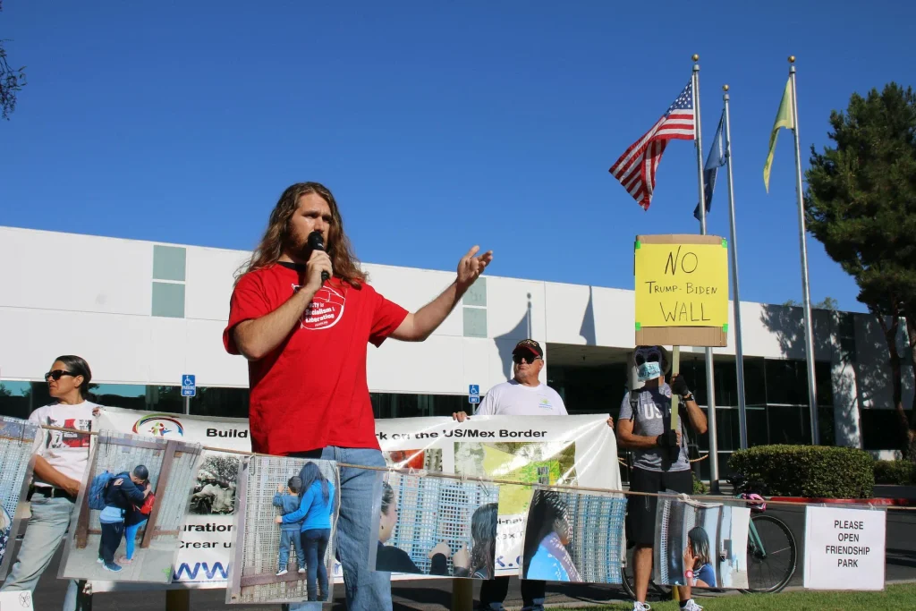 A man with long hair speaks into a microphone at an outdoor protest. Behind him, people hold banners and a yellow sign reading "No Trump-Biden Wall." U.S. flags and a modern building are visible in the background.