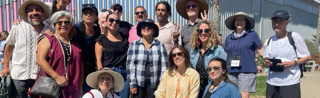 A group of smiling adults poses outdoors in front of a colorful corrugated metal wall. Most are wearing hats and sunglasses, and the weather appears bright and sunny.