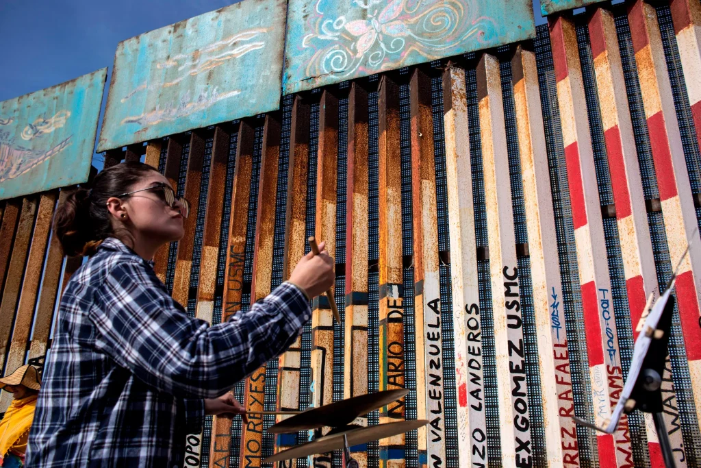 A woman wearing sunglasses and a plaid shirt plays cymbals in front of a rusted border fence painted with names and colorful artwork under a blue sky.