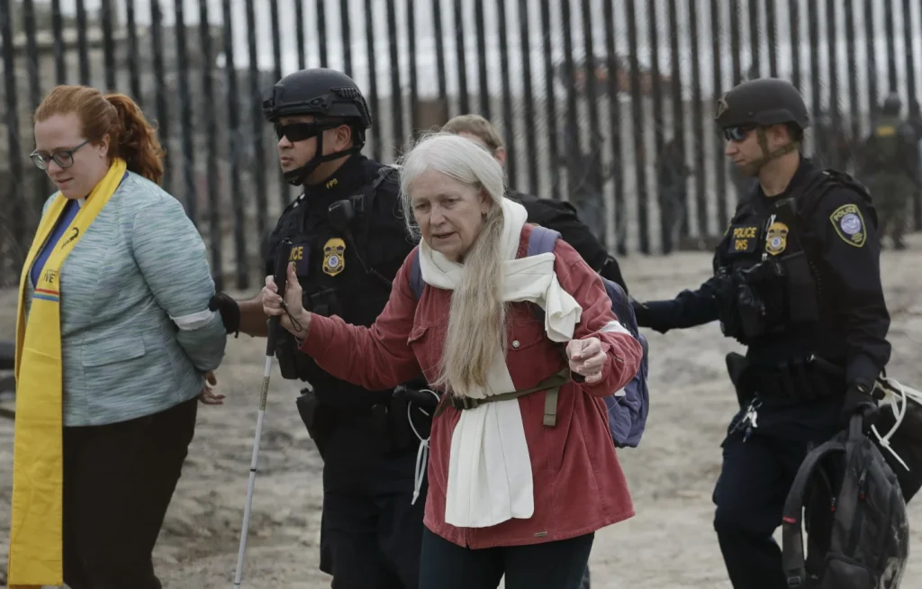 Two women are being escorted by police officers in tactical gear near a border fence. One woman has long white hair and wears a red jacket, while another wears glasses and a yellow scarf. The background is a sandy outdoor area.